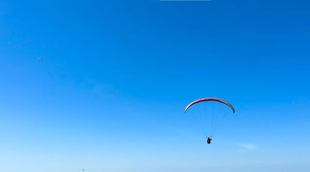Paraglider in flight above the Bir landing ground, with hills and houses below