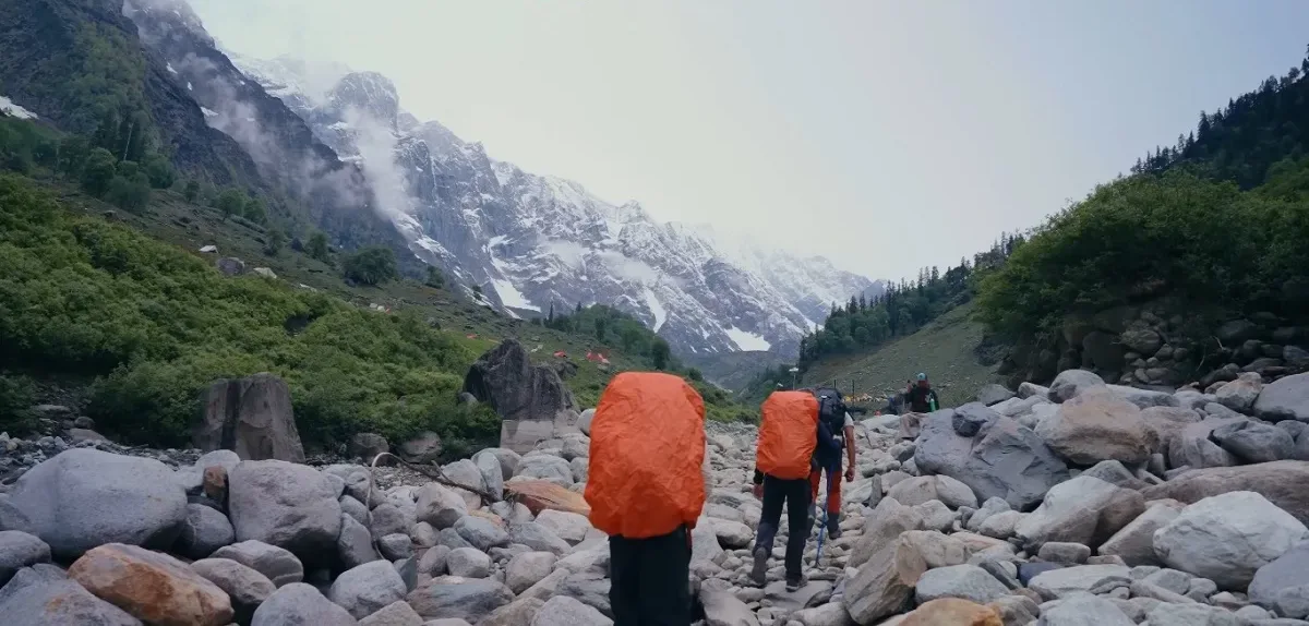 Trekkers crossing a boulder-filled valley towards a riverside campsite below snow-capped mountains on the Friendship Peak trek
