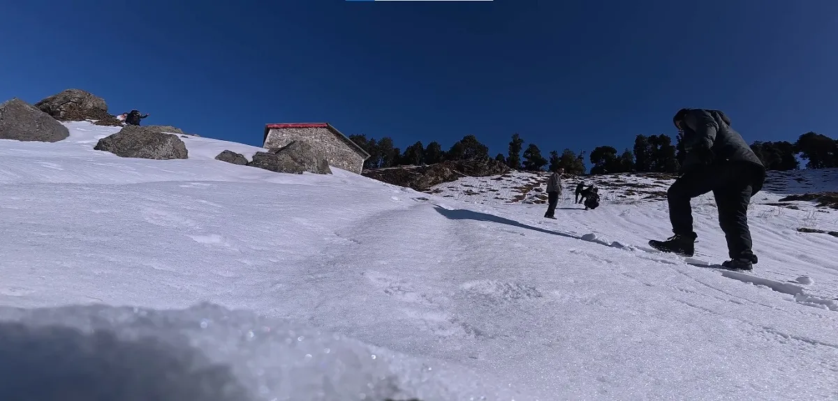 Trekkers climbing a snow-covered slope towards the stone hut below Nag Tibba summit